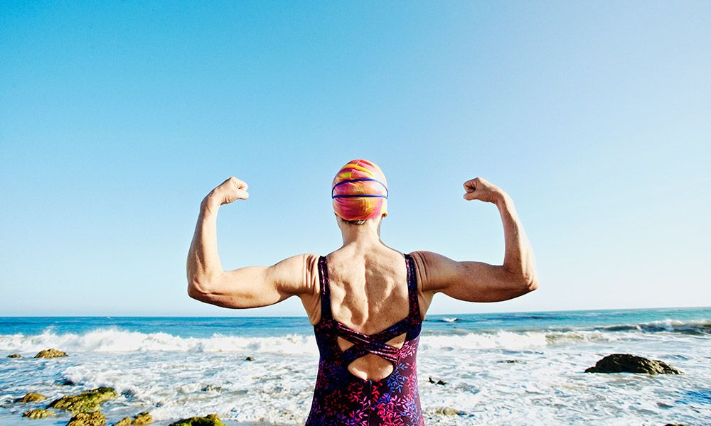 older woman in a swimsuit and cap flexing her muscles at the beach.
